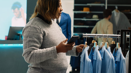 Female client checking online website stock in shop, trying to find formal wear from internet app in department store. Young adult using phone to buy favorite clothes in shopping center.の写真素材