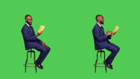 Young businessman sitting on chair reading story book or novel, being focused on fictional tale for leisure activity. Confident entrepreneur reading literature over full body green screen.の写真素材