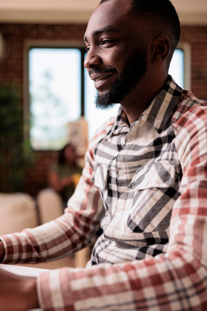 Portrait of smiling african american man browsing social media content on computer screen while girlfriend is reading a book on the couch. Freelancer working remote sitting at desk in home living room.の写真素材