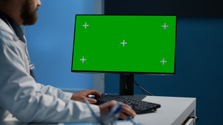 Doctor typing medical expertise on computer with green screen chroma key display working over hours in hospital office. Practitioner medic checking patient report while planning health care treatmentの写真素材