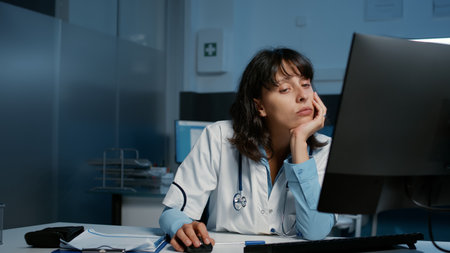 Exhausted stressed practitioner doctor working late at night in hospital office typing patient disease expertise while falling asleep on desk. Tired frustrated medic checking medical report on computerの写真素材
