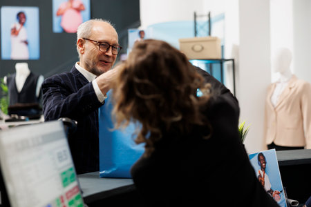 Old client taking shopping bag from boutique employee, after buying fashionable clothes in shopping centre. Senior shopper paying for stylish merchandise during commercial activityの写真素材