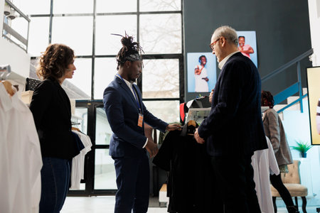 Elderly customer wanting to buy formal suit, asking showroom employee for help to choose the right clothes. Senior man buying fashionable merchandise and stylish accessories in clothing storeの写真素材