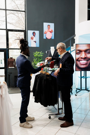 Elderly customer discussing merchandise fabric with worker, looking to buy formal wear in clothing store. Senior client buying fashionable clothes to increase wardrobe in showroom. Fashion conceptの写真素材