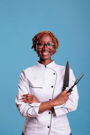 African american female cooker, curly haired posing with professional kitchen knife, sharpener. Smiling afro chef holding kitchen utensils. Portrait of uniformed young female restaurant worker.の写真素材