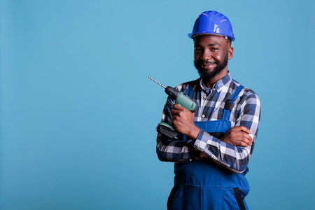 African american builder holding electric cordless drill posing happily in studio. Constructor wearing hard hat and construction coverall, safety uniform in construction industry.の写真素材