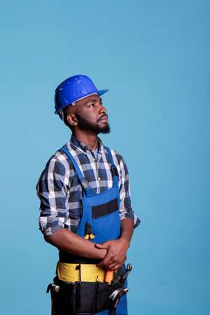 African american builder looking intently up, detailing the tools and materials needed to do the job. Construction worker wearing coveralls and hard hat in studio shot against blue background.の写真素材