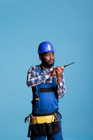 Serious builder screwing something into construction with concentrated expression, wearing blue overalls and protective helmet. Interior studio shot isolated on blue background.の写真素材