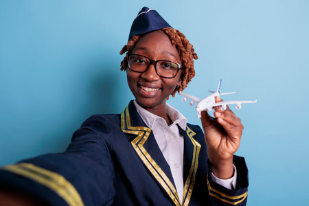 Smiling female flight attendant in uniform holding and playing with model airplane. Aircraft crew member looking at commercial jet toy in studio shot against blue background.の写真素材