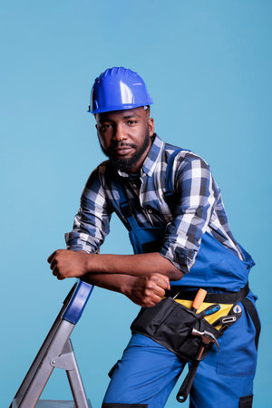 Builder looking calm leaning against ladder in studio shot against blue background. African american construction worker wearing hard hat and work uniform looking into camera.の写真素材