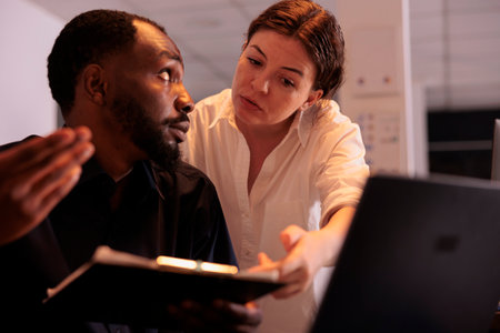 Analyst helping colleague analyzing report statistics, comparing data on laptop in office. Two corporate workers checking marketing research results in coworking space at nightの写真素材