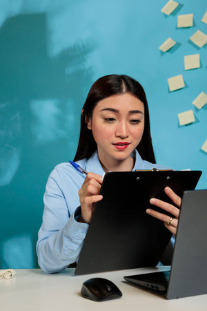 Employee working from office looking at paperwork on clipboard attentive to task list. Professional woman reviewing notes while working on important project doing overtime.の写真素材