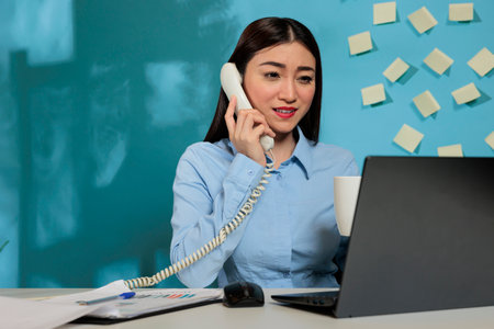 Asian woman talking on the landline phone and smiling while sitting at her working place in the...