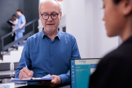 Elderly patient standing at hospital reception signing medical insurance paper while discussing medication treatment with receptionist. Old man having checkup appointment in hospital lobbyの写真素材
