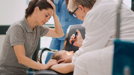 Diverse medical team consulting little girl being unconscious, using stethoscope and flashlight in facility lobby. Sick child fainting on chairs and worried mother trying to help. Handheld shot.の写真素材