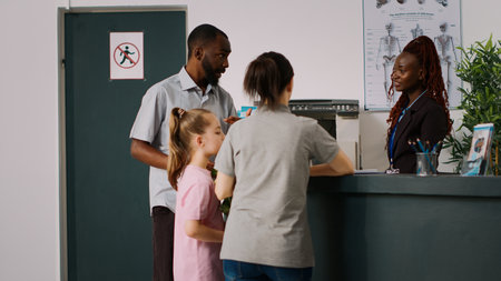 Young family asking receptionist about appointment with medic, sitting at reception counter and filling in checkup report. Man and woman with child talking to medical worker in lobby.の写真素材