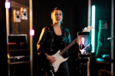 Guitarist performer holding electric guitar playing heavy metal song while recording performance with phone during music session in audio studio. Woman musician working at new grunge album before tourの写真素材