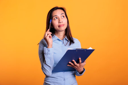 Pensive woman thinking at ideas for business project, writing report on documents. Thoughtful filipino adult looking at checklist, answering questionnaire in studio over yellow backgroundの写真素材
