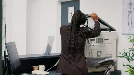 Medical employee working at hospital reception desk, using landline phone to make checkup appointments with patients. Facility worker planning healthcare consultations at counter. Handheld shot.の写真素材
