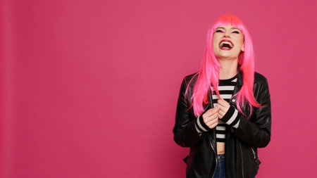 Cheerful lovely woman with pink hair wig laughing and having fun on camera, showing candid smile in studio. Young happy girl with leather jacket enjoying jokes and having laugh.の写真素材