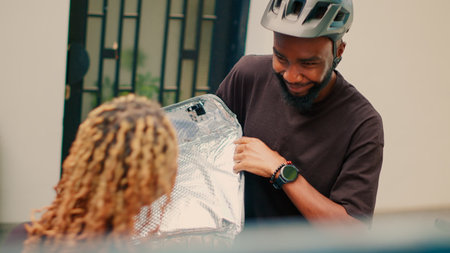 Express meal delivery service delievered by employee at building entrance, giving client paperbag order. Female client holding fastfood takeaway meal in package, delivering on bike.の写真素材
