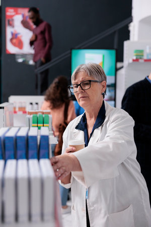 Elderly pharmacist in white coat looking at pills packages checking pharmacy shelves while helping people with health care treatment. Diverse customer buying vitamins, supplements, health care serviceの写真素材