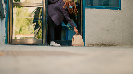 Female carrier letting fastfood order package at front door, client picking up paperbag with restaurant lunch. Takeaway service courier delivering food during covid 19 pandemic, wearing facemask.の写真素材