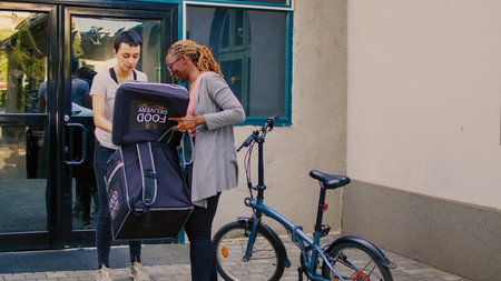 Food delivery courier giving meal in paperbag to woman, delivering restaurant food on bicycle. Riding bike and carrying backpack to deliver takeaway fastfood package at building entrance.の写真素材