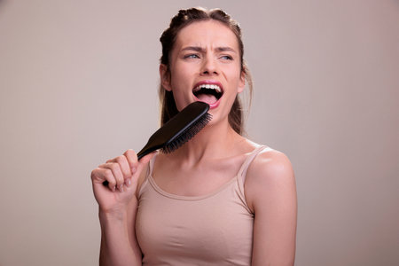 Young beautiful woman holding hairbrush pretending to be singer and using comb as microphone. Blonde lady wearing beige casual top brushing hair, listening to music and singingの写真素材
