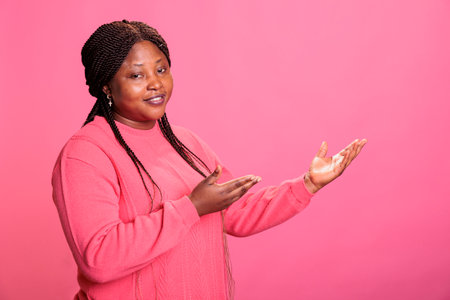 Portrait of attractive young adult advertising promotional product in studio, posing for advertisement over pink background. Confident woman with stylish hairstyle looking at camera during ad shootの写真素材