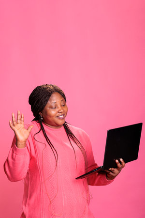 Smiling young adult holding laptop computer greeting remote friend during online videocall meeting conference, enjoying discussion while standing over pink background. Model with positivity emotionの写真素材
