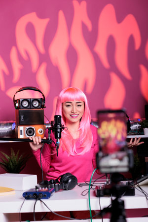 Asian woman with pink hair talking with subscribers using professional microphone while filming broadcast in vlogging studio. Influencer recording podcast with mobile phone camera, live streamingの写真素材