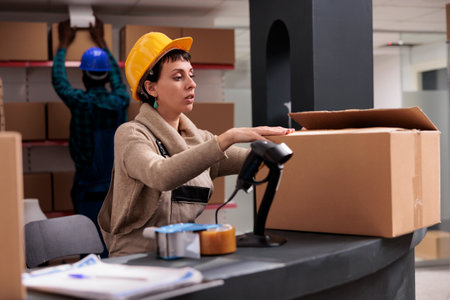 Retail storehouse manager packing cardboard box at counter desk and closing package. Caucasian warehouse worker wearing safety helmet preparing customer parcel for deliveryの写真素材