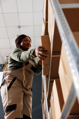 Stockroom employee taking out cardboard box from metallic shelf, preparing clients orders in storage...