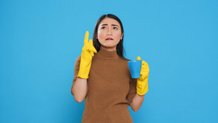 Pensive thoughtful maid holding cup of coffee standing over blue background in studio, thinking at solution to solve cleaning problem. Housekepper providing top-notch cleaning services to her clients.の写真素材