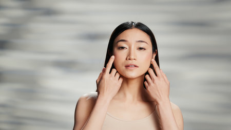 Flawless woman with radiant skin promoting beauty ad, with products for luminous glowing appearance. Asian girl posing on camera and advertising empowering skincare campaign.の写真素材
