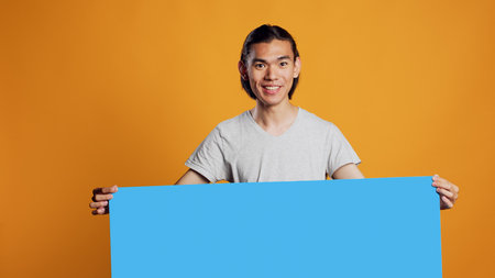 Asian young man holding blue carton board on camera, using isolated copyspace on billboard in...