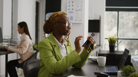 African american employee using mobile phone at work, sitting at desk planning business investment. Young manager checking social media app on smartphone, career development.の写真素材