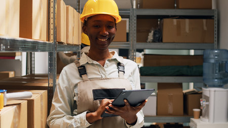 Female employee in overalls checking tablet inventory, using device to plan logistics to ship...