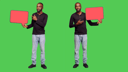 Male model holding red blank speech bubble on camera, creating presentation with empty copyspace cardboard icon. Young adult showing mockup billboard over green screen backdrop.の写真素材