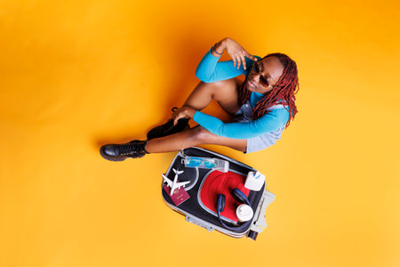 African american girl with sunglasses, posing in studio on the floor with suitcase and items. Cheerful person having trolley bag with multiple essentials and objects, travelling on vacation.の写真素材