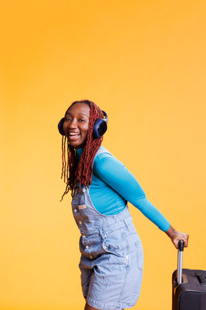 Female tourist listening music on trip, carrying big trolley bag for weekend adventure or modern city break. Cheerful woman wearing headphones on airplane flight, smiling in studio.の写真素材