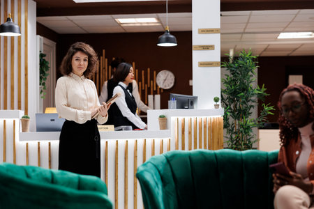 Portrait of hotel manager with records standing in reception desk lobby area to check concierge service as supervisor. Female lodging manager and administrator welcoming guests.の写真素材