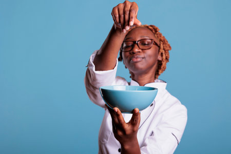 African American chef holding bowl of food while adding salt with style in front of the camera. Professional cook proud of new preparation in studio shot against blue background.の写真素材