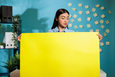 Optimistic asian woman holding yellow cardboard sign while posing in modern office on blue background. Confident female employee showing advertising poster with copy space.の写真素材
