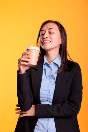 Satisfied asian model holding cup of coffee drinking hot beverage enjoying free time before start to photoshot in studio. cheerful young adult with brunette long hair standing over yellow backgroundの写真素材