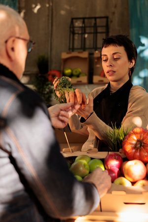 Young woman farmer standing behind farmers market stall showing organic locally grown carrots to customer while selling fresh fruits and vegetables. Mature man buying healthy local foodの写真素材