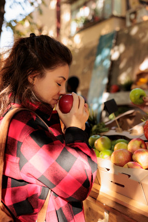 Female client smelling apples and shopping at farmers market, buyer standing near market stall choosing locally grown fresh organic fruits and vegetables. Visiting harvest fair festival.の写真素材