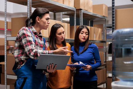 Warehouse employees team cooperating and preparing order using pick ticket on laptop. Storehouse asian workers doing inventory management and solving delivery problems togetherの写真素材