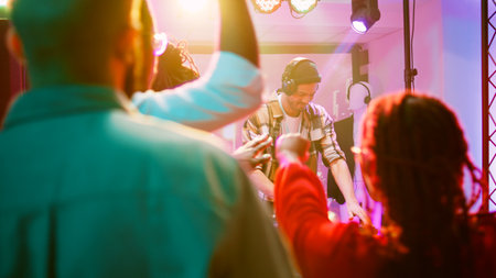 Persons dancing with DJ at nightclub, young adult mixing funky music for group of friends on dance floor. Happy people partying and having fun at celebration with disco lights. Handheld shot.の写真素材